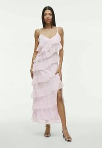 Lang rosa kjole med lagdelte volanger, tynne stropper og en høy slitt opp til låret, laget av lett materiale. Sølvfargede hælsandaler kompletterer antrekket.