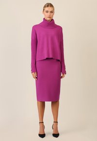 Suéter fucsia con un cuello alto y mangas acanaladas, combinado con una falda lápiz a la altura de la rodilla. Ambos hechos de tela de punto suave. Zapatos de tacón negros.