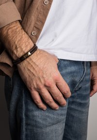 Rudas pintas odinė apyrankė su auksiniu magnetiniu užsegimu, dėvima ant rankos, restingant ant mėlynų džinsų, neutralioje fone.