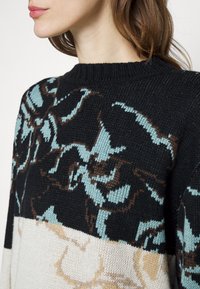 Pull en tricot avec un motif floral en noir, blanc et pastel, col rond côtelé et tissu texturé. Contrastes de couleurs subtils.