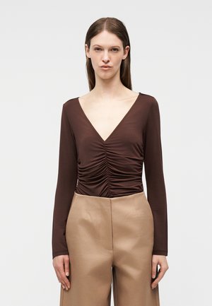 Kobieta o prostych brązowych włosach, ubrana w brązową marszczoną bluzkę z długim rękawem i dekoltem w kształcie litery V oraz beżowe, wysokowatowe spodnie z szerokimi nogawkami, stojąca na tle białego tła.