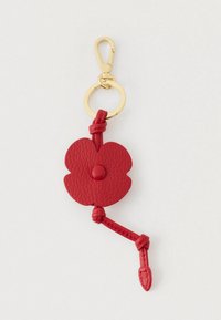 Zelta atslēgu piekariņš ar sarkano ādas zieda formas piekariņu, kas rotāts ar mezglota ādas siksniņu uz balta fona.