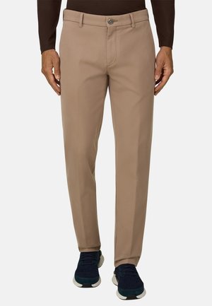 Homme portant un pantalon beige droit et des baskets foncées, debout les mains relâchées le long du corps devant un fond uni.