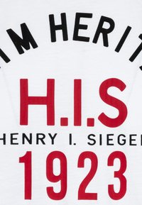 Camiseta blanca con texto en negro y rojo. El texto incluye "H.I.S," "HENRY I. SIEGER," y "1923." Diseño simple con una textura suave.