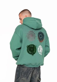 Grøn hættebluse med løs pasform, der har fire grafiske tryk på ryggen i sort og grøn samt et textured stof.