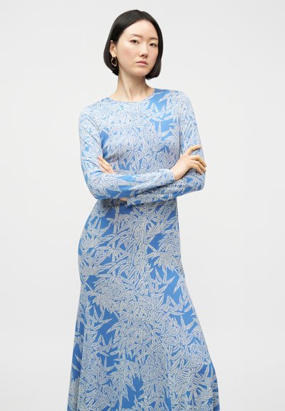 Γυναίκα με κοντά μαύρα μαλλιά, φοράει μπλε φόρεμα με μακριά μανίκια και λευκό μοτίβο φύλλων, με σταυρωμένα χέρια, με ουδέτερη έκφραση, και απλό φωτεινό φόντο.