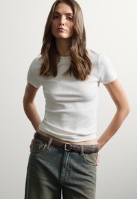 Top blanco de manga corta con diseño ajustado combinado con jeans azules desgastados de cintura alta y un cinturón de cuero texturizado marrón.
