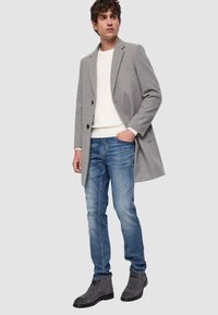 Cappotto grigio in mélange di lana con due bottoni, indossato sopra un maglione bianco testurizzato e jeans blu, abbinato a stivaletti grigi alla caviglia.