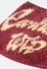 Tessuto marrone scuro con una texture fuzzy che presenta lettere gialle. Il design include una forma circolare e cuciture visibili lungo il bordo.