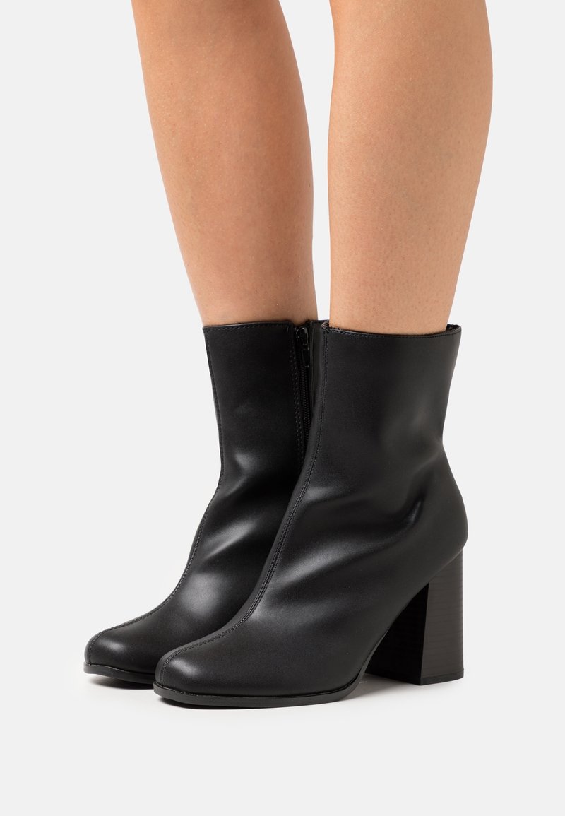 Bottes en cuir noir montant à la cheville avec des talons épais portées sur des jambes nues sur un fond clair uni.
