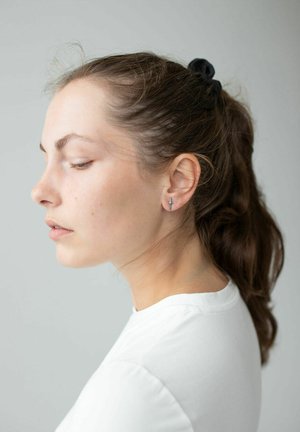 Silver ear stud featuring a minimalist sword design, worn by a person with long, dark hair tied back, dressed in a white crew-neck shirt.