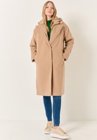 Cappotto lungo beige con cappuccio, realizzato in un tessuto morbido e strutturato e dalla vestibilità ampia. Indossato sopra un maglione verde e abbinato a jeans blu e sneakers beige.