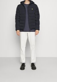 Marineblaue Steppjacke mit Kapuze, gestepptem Design und kleinem gelbem Logo; getragen über einem marineblauen, strukturierten Hemd und weißen Hosen mit schwarzen Schuhen.