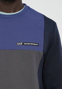 Tamno plavi dukserica s sivim i ljubičastim dijelovima, s okruglim izrezom i oznakom EA7 Emporio Armani na prsima.