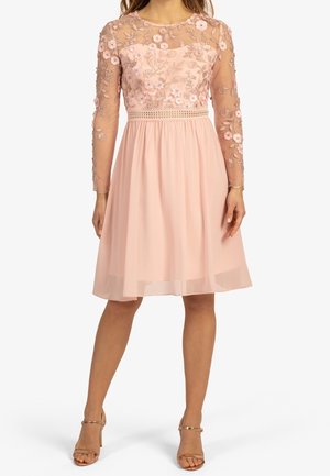 Frau trägt ein knielanges pinkes Kleid mit transparenten, langärmligen Ärmeln mit Blumenstickerei und taillierter Passform, kombiniert mit goldenen Riemchen-Absätzen.