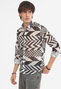 Camicia a maniche lunghe con bottoni in bianco e nero, caratterizzata da un motivo geometrico, realizzata in un tessuto leggero e semi-trasparente. Maniche lunghe arrotolate.