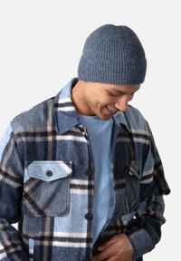 Chemise en laine à carreaux bleus avec deux poches poitrine, associée à un t-shirt bleu clair et un bonnet en maille grise texturée.