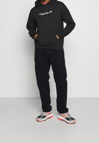 Sudadera negra con texto blanco, combinada con pantalones oscuros y zapatillas deportivas multicolores con acentos en gris, naranja y rosa.