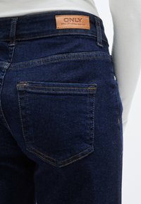 Σκούρο μπλε τζιν με ψηλή μέση, με πορτοκαλί ραφές, δύο πίσω τσέπες και καφέ δερμάτινη ετικέτα με το "ONLY" στη μέση.