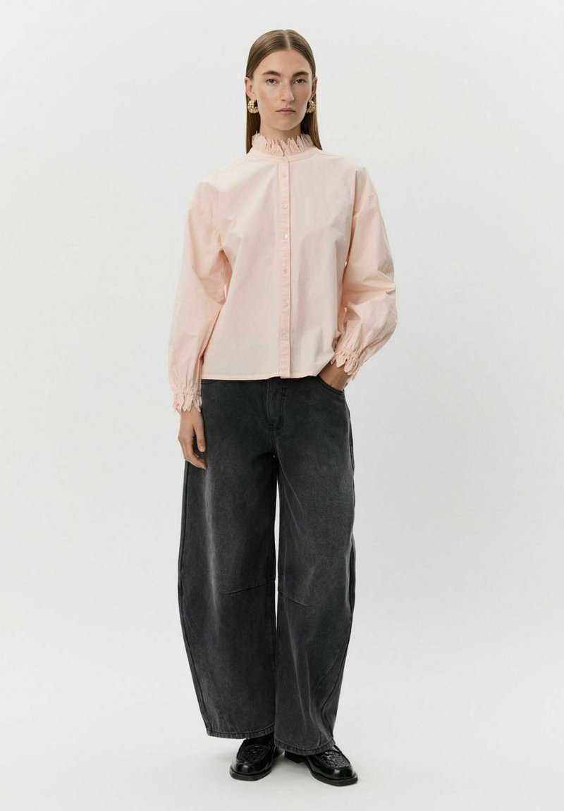 Svetloružová košeľa s golierikom a balónovými rukávmi so stiahnutými manžetami, v kombinácii s voľnými čiernymi džínsami a čiernymi topánkami.