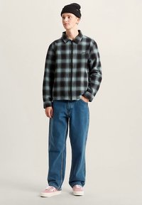 Giacca di flanella a quadri neri e blu con cerniera e collo con risvolto, abbinata a jeans di denim blu larghi e sneakers rosa chiaro.