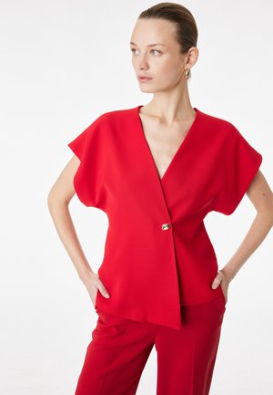 Kvinde iført en rød V-hals top med brede korte ærmer og en enkelt knap, kombineret med matchende røde bukser, stående med hænderne på hofterne.