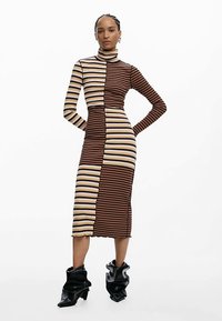 Robe en maille avec un col montant, présentant des rayures horizontales alternées marron et beige, une silhouette ajustée et une texture côtelée. Portée avec des bottes noires.