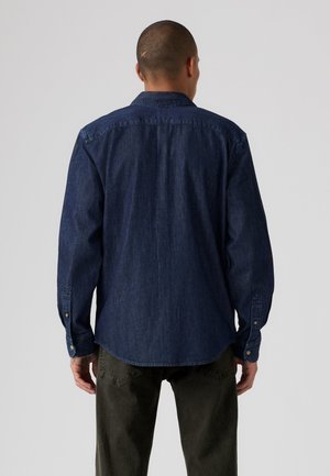 Homme debout de dos, portant une chemise en jean bleu foncé à manches longues et un pantalon vert foncé sur un fond clair uni.