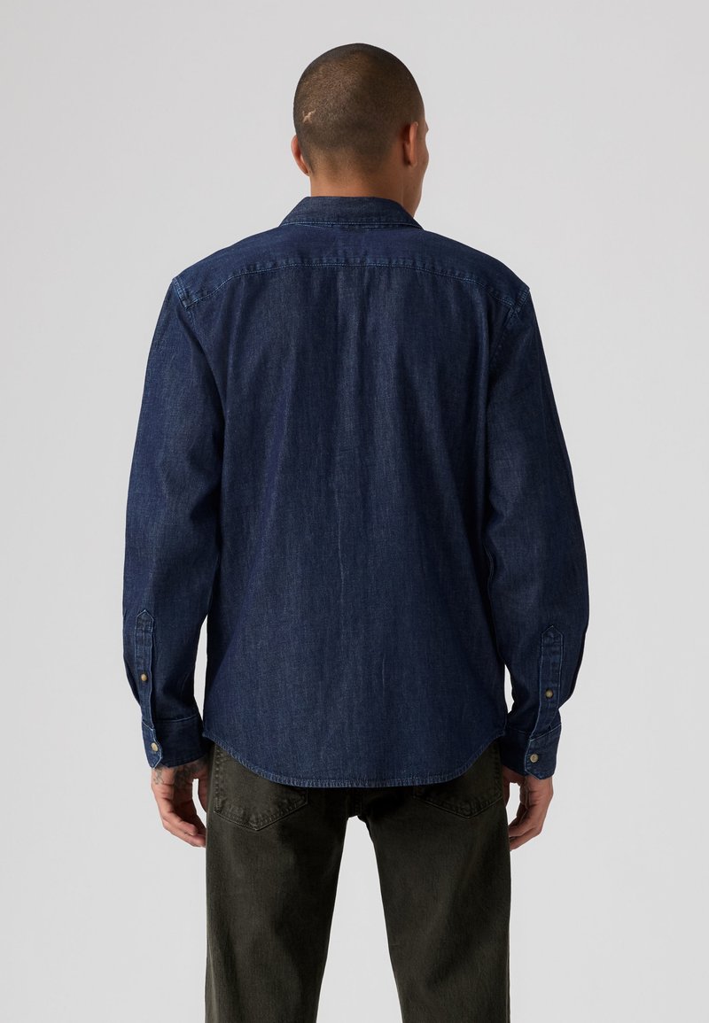 Homme debout de dos, portant une chemise en jean bleu foncé à manches longues et un pantalon vert foncé sur un fond clair uni.