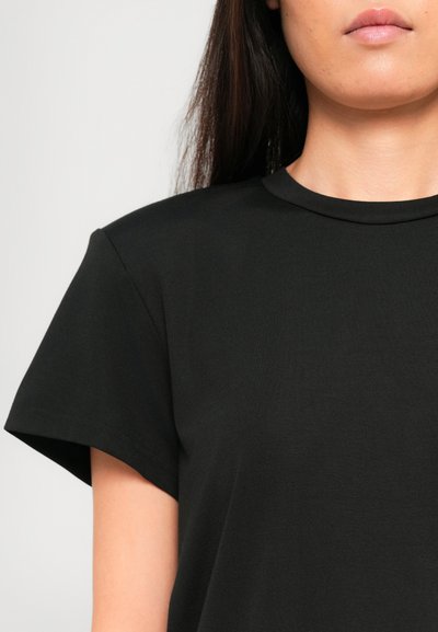 Woman wearing a plain black short-sleeve t-shirt, showing upper torso and part of the face against a light background.