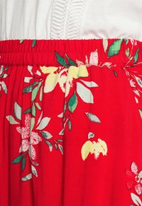 Red fabric with floral patterns in yellow and green; features an elastic waistband and pleats for added texture and detail.