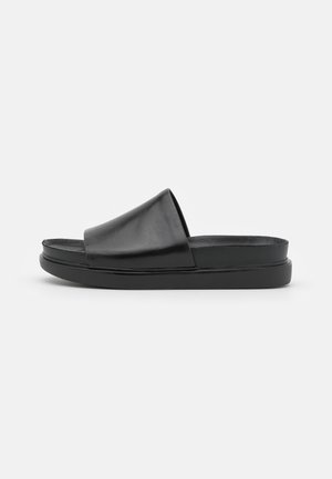 Sort lædersandal med åben tå, der har en bred rem, flad sål og minimalistisk design. Overdelen har en glat tekstur og en rund form.
