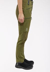 Olivově zelené outdoorové kalhoty se zipovými kapsami, zesílenými detaily a slim fit. S černými turistickými botami. Logo je viditelné na obou.