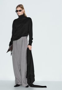 Maglione nero a collo alto, pantaloni grigi a gamba larga e accessori neri minimal. Texture eleganti e lisce con pieghe e drappeggi pronunciate.