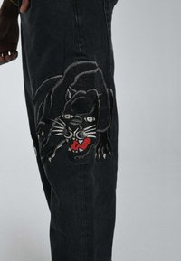 Pantalones vaqueros negros con un gran diseño bordado de pantera en la pierna izquierda, con acentos rojos para la boca y rasgos faciales detallados.