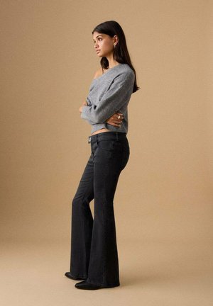 Grå strikket sweater, mørke højhalset flare-jeans, sorte ankelstøvler, ringe på fingrene, stående i profil mod en beige baggrund.