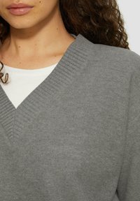Grauer V-Ausschnitt-Pullover aus weichem Strickstoff mit gerippten Akzenten am Kragen und einer darunter sichtbaren weißen Innenschicht.