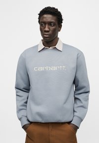 Άνδρας που φοράει ανοιχτό μπλε φούτερ Carhartt πάνω από μπεζ πουκάμισο με γιακά και καφέ παντελόνι, με τα χέρια στις τσέπες, κοιτάζει στο πλάι μπροστά σε λευκό φόντο.