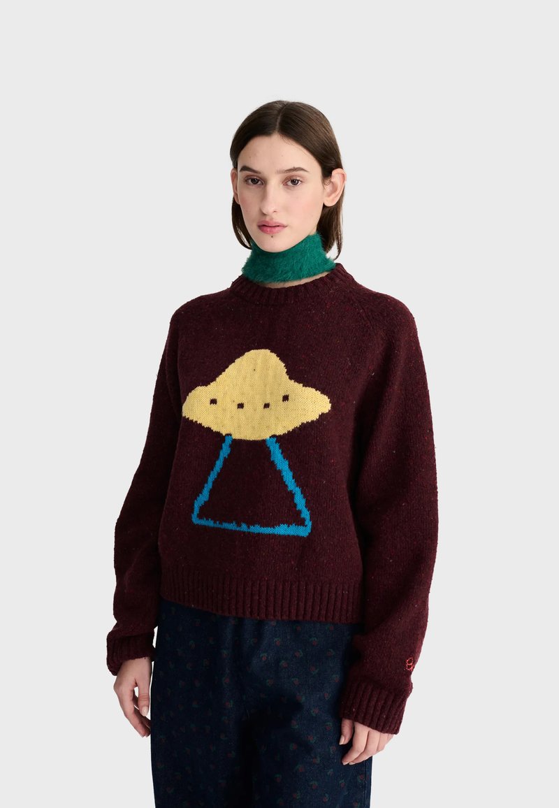 Jeune femme portant un pull rouge foncé avec un motif OVNI jaune et bleu et un col roulé vert sous le pull.