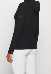 Felpa nera con cappuccio in tessuto testurizzato, caratterizzata da un design aderente, maniche lunghe e un accento del logo Nike dorato sul braccio.