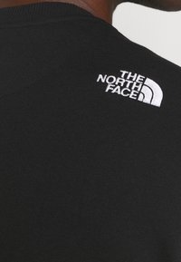Svart tyg t-shirt med rund halsringning, med en vit broderad logo av The North Face på vänster axel. Slät textur.