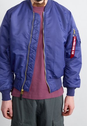 Homme portant une veste bomber bleue avec une étiquette "Remove Before Flight" sur la poche de la manche, une chemise bordeaux et un pantalon vert foncé.