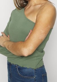 Olive green, ribbed sweater with an asymmetric neckline and one long sleeve; hemline is scalloped. Worn with blue denim jeans.