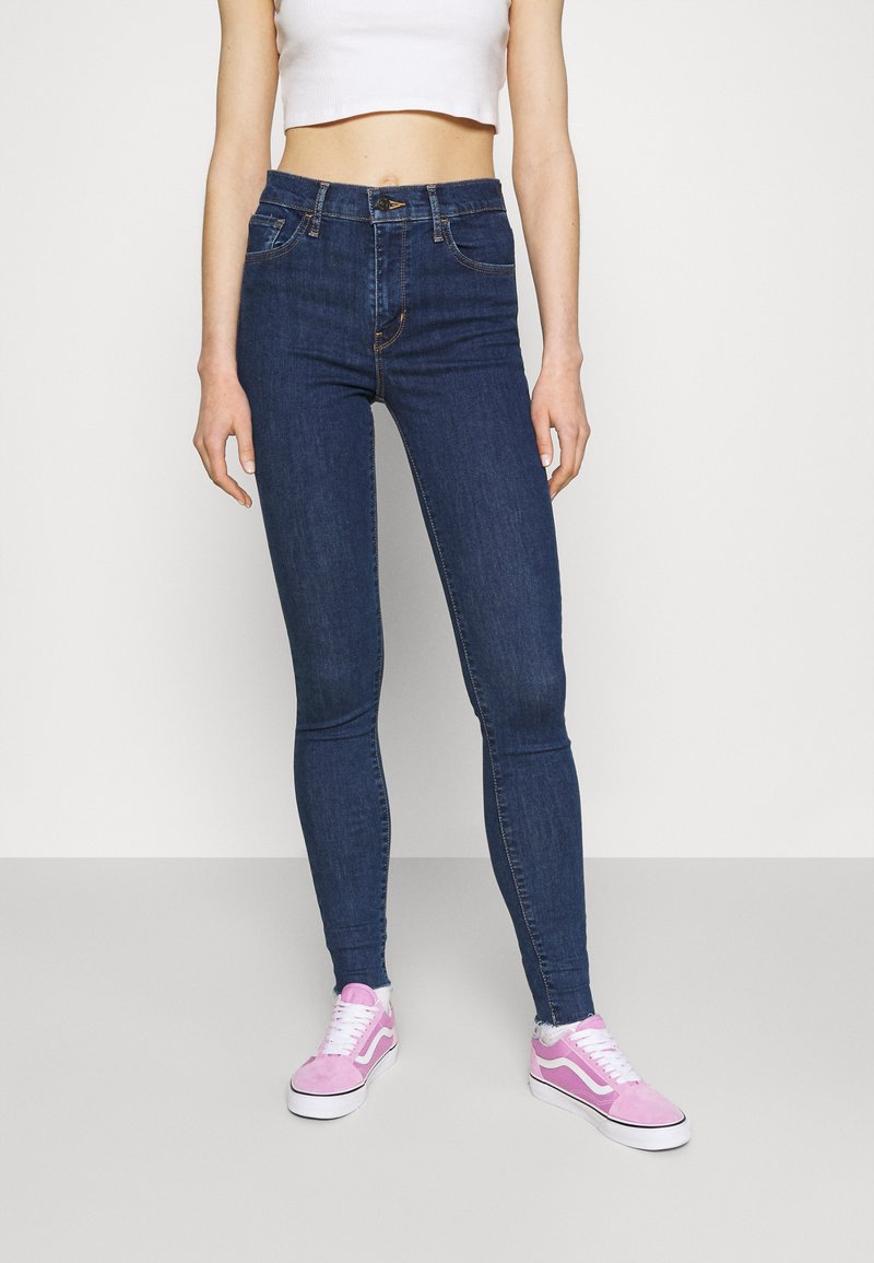Jeans skinny feitos de denim azul escuro com uma cintura alta, apresentando cinco bolsos e costura laranja, combinados com ténis de camurça cor-de-rosa.