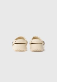 Beige Crocs mit einem Riemen an der Rückseite, aus leichtem Material und mit strukturiertem Fußbett. Das Wort "Crocs" ist auf dem Fersenriemen eingraviert.