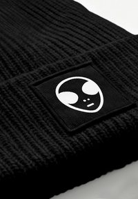Black knit beanie featuring a square patch with a white alien face design. The texture is ribbed, providing stretch and warmth.