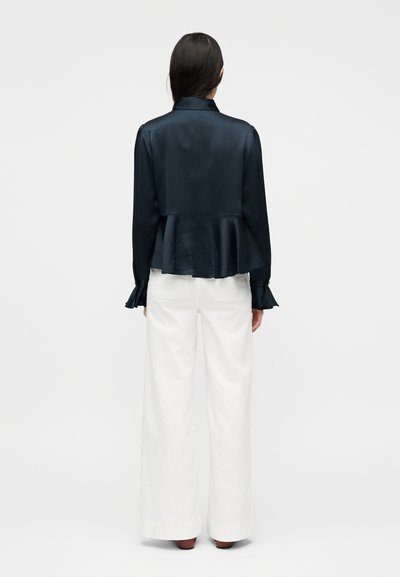 Γυναίκα φτιαγμένη από πίσω που φοράει μαύρη σατέν μπλούζα με πεπλούμ, βολάν στα μανίκια και πλατύχωρα λευκά παντελόνια, πάνω σε απλό φόντο.