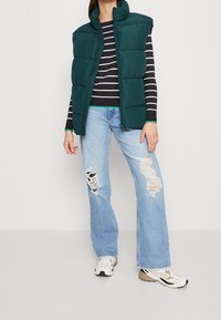 Grüne Steppweste mit hohem Kragen, über einem gestreiften schwarz-weißen Langarmshirt getragen. Kombiniert mit hellblauen, verwaschenen Jeans und Sneakers.