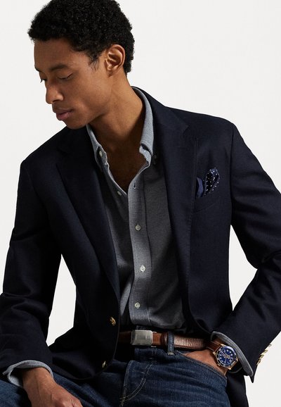Navy blazer over a gray button-up shirt, paired with dark jeans and a brown belt. Notable details include a pocket square and a blue watch.