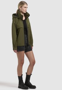 Veste à capuche vert olive, fermeture éclair, deux poches sur la poitrine, portée sur un haut noir et un short noir, associée à des bottes noires jusqu'aux genoux.